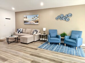 Ein Wohnzimmer mit einem beigen Sofa, zwei blauen Sesseln, einem Couchtisch, einem gemusterten Teppich, einem Bild von einer Strandlandschaft und einer Wanddekoration mit kreisförmigen Formen.