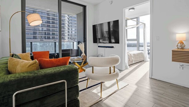 A modern living room with a green sofa, a white chair, a flat-screen TV, and a large window overlooking a cityscape. - Downtown Miami 2BR Skyline Bay View Balcony Corner Suite Luxepad (Miami)