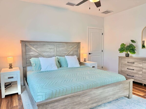 A bedroom with a wooden headboard, a bed with a light green quilt, two bedside tables with lamps, a wooden dresser, and a potted plant. - The Stingray Glide Into Your Beach Adventure! (Punta Gorda)