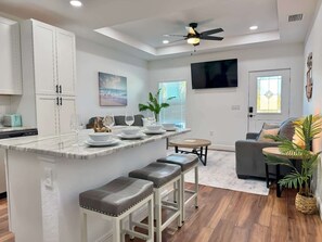 A modern kitchen with a marble island, bar stools, a dining area with a round table, and a living room with a sofa and a TV. - The Stingray Glide Into Your Beach Adventure! (Punta Gorda)
