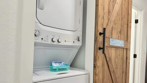 A laundry area with a white washing machine, a wooden door with a sign reading "LAUNDRY SELF SERVICE OPEN 24 HOURS," and a basket with a Snuggle detergent box. - The Stingray Glide Into Your Beach Adventure! (Punta Gorda)