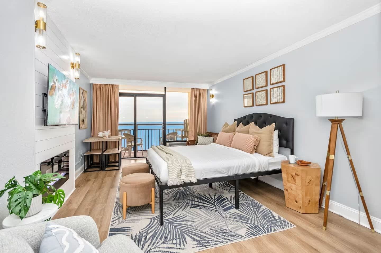 A hotel room with a bed, a desk, a chair, a small table, a potted plant, a lamp, and a view of the sea.