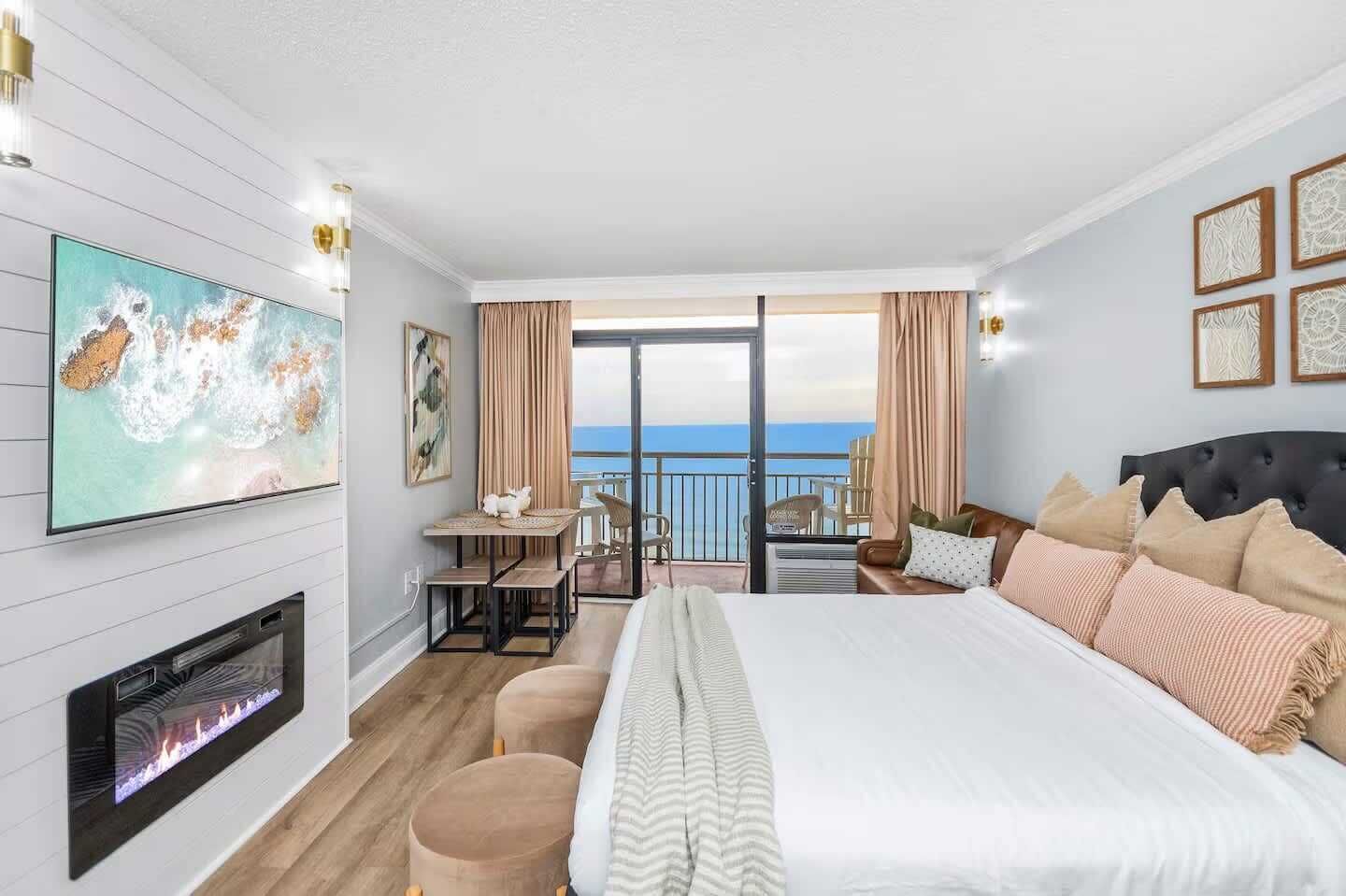 A hotel room with a bed, a fireplace, a balcony with a view, and a dining area.