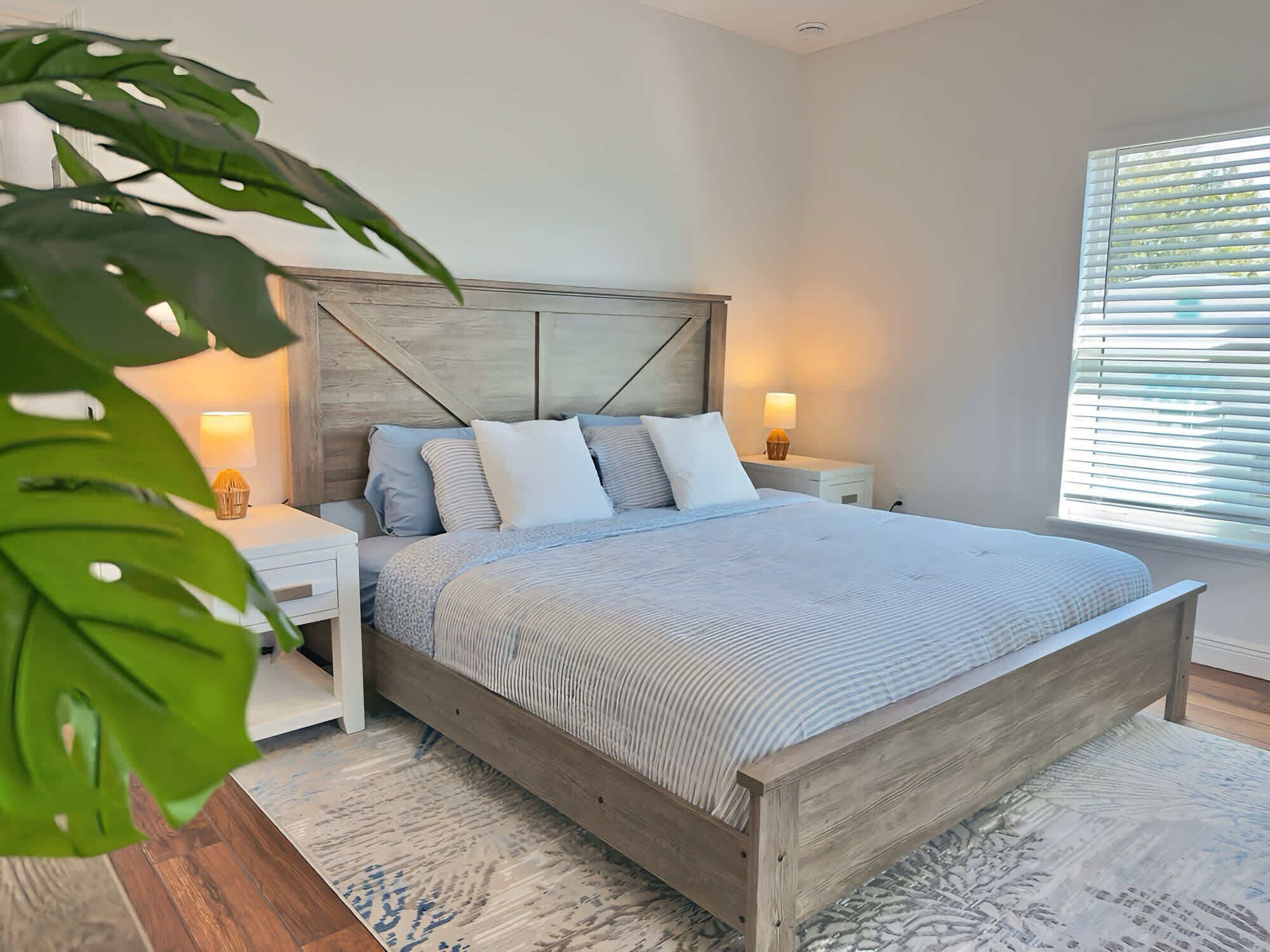 A bedroom with a wooden bed frame, a nightstand with a lamp, and a large window with blinds.