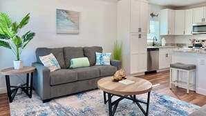 Een modern appartement met een grijze bank, een ronde salontafel en een keuken met witte kasten en een magnetron.