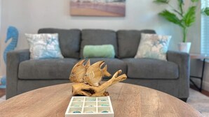 A living room with a grey sofa, decorative pillows, a wooden table with a fish sculpture, and a painting of a beach scene on the wall. - Seahorse Drift Away in Coastal Comfort (Punta Gorda)