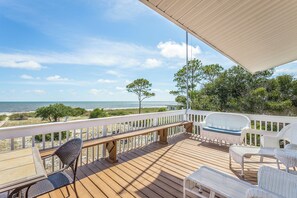 Eine Holzterrasse mit weißen Korbmöbeln, einer Bank und Blick auf Strand und Meer.