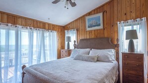 Een slaapkamer met een houten lambrisering, een bed met witte lakens, een plafondventilator en een raam met transparante gordijnen.