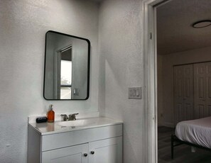 A bathroom with a white vanity, a mirror, a sink, and a toilet. There is a door leading to another room. - Cozy 2br Apartment in Cape Coral With Hot Tub U201 (Cape Coral)