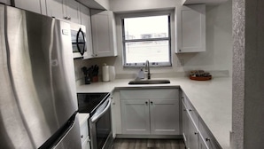 A compact kitchen with white cabinets, a stainless steel refrigerator, a built-in oven, a window with blinds, and a sink with a soap dispenser. - Cozy 2br Apartment in Cape Coral With Hot Tub U201 (Cape Coral)