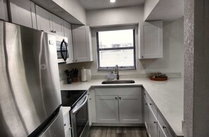 A compact kitchen with white cabinets, a stainless steel refrigerator, a built-in oven, a window with blinds, and a sink with a soap dispenser. - Cozy 2br Apartment in Cape Coral With Hot Tub U201 (Cape Coral)
