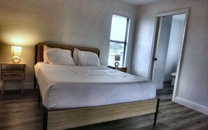 A bedroom with a bed, a nightstand, a lamp, a window, and a door leading to a bathroom. - Cozy 2br Apartment in Cape Coral With Hot Tub U201 (Cape Coral)