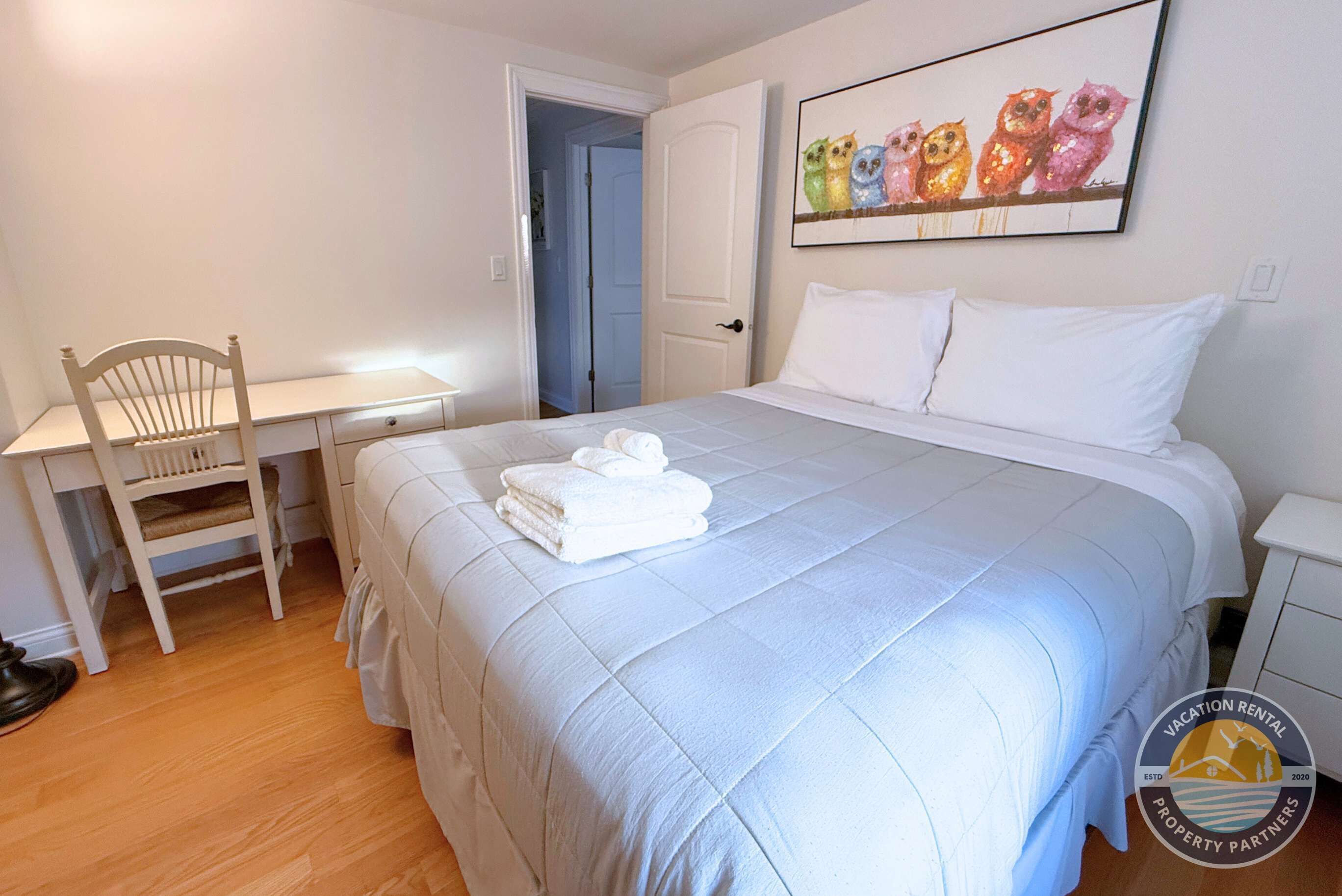 A bedroom with a bed, a desk, and a chair. There is a painting on the wall and a logo on the bed.