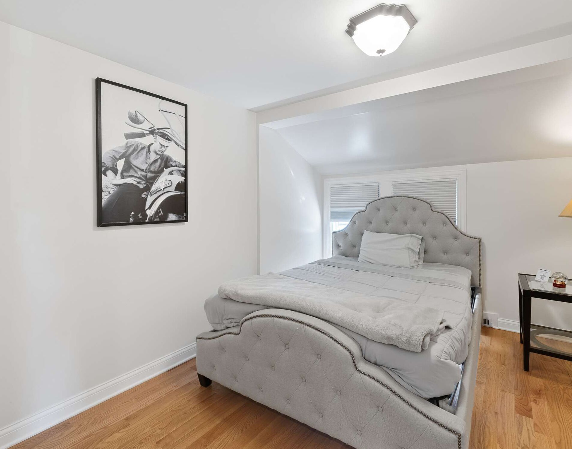 A bedroom with a tufted headboard bed, a framed black and white photograph on the wall, and a small bedside table with a lamp.