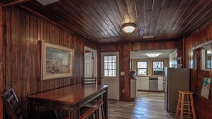 Um quarto com painéis de madeira, uma mesa de jantar e cadeiras, uma área de cozinha com eletrodomésticos e uma moldura com um quadro na parede.