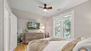 A bedroom with a bed, a ceiling fan, a dresser, a chair, and a window with a view of trees. - Walk to Lake Relaxing Williams Bay Getaway (Williams Bay)