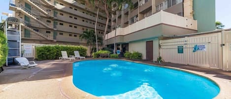 A multi-story building with a swimming pool, lounge chairs, and a gated entrance.