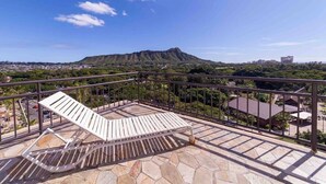 A balcony with a white lounge chair, stone flooring, and a view of a mountain and cityscape. - Marbella Lane - Waikiki Studio Beach 15 Steps (Honolulu)