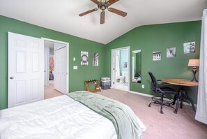 Un dormitorio con cama, escritorio y silla, ventilador de techo, y baño visible a través de una puerta abierta.
