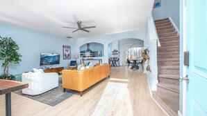 A spacious living room with a large TV, a sofa, and a dining area with a table and chairs. There is a staircase leading to another level. - Modern 3-br Townhome w Private Backyard Near RTP (Morrisville)