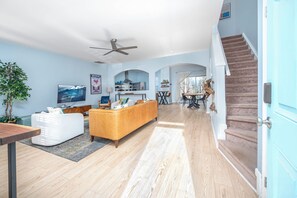 Amplia sala de estar con televisor grande, sofá y comedor con mesa y sillas. Una escalera conduce a otro nivel.