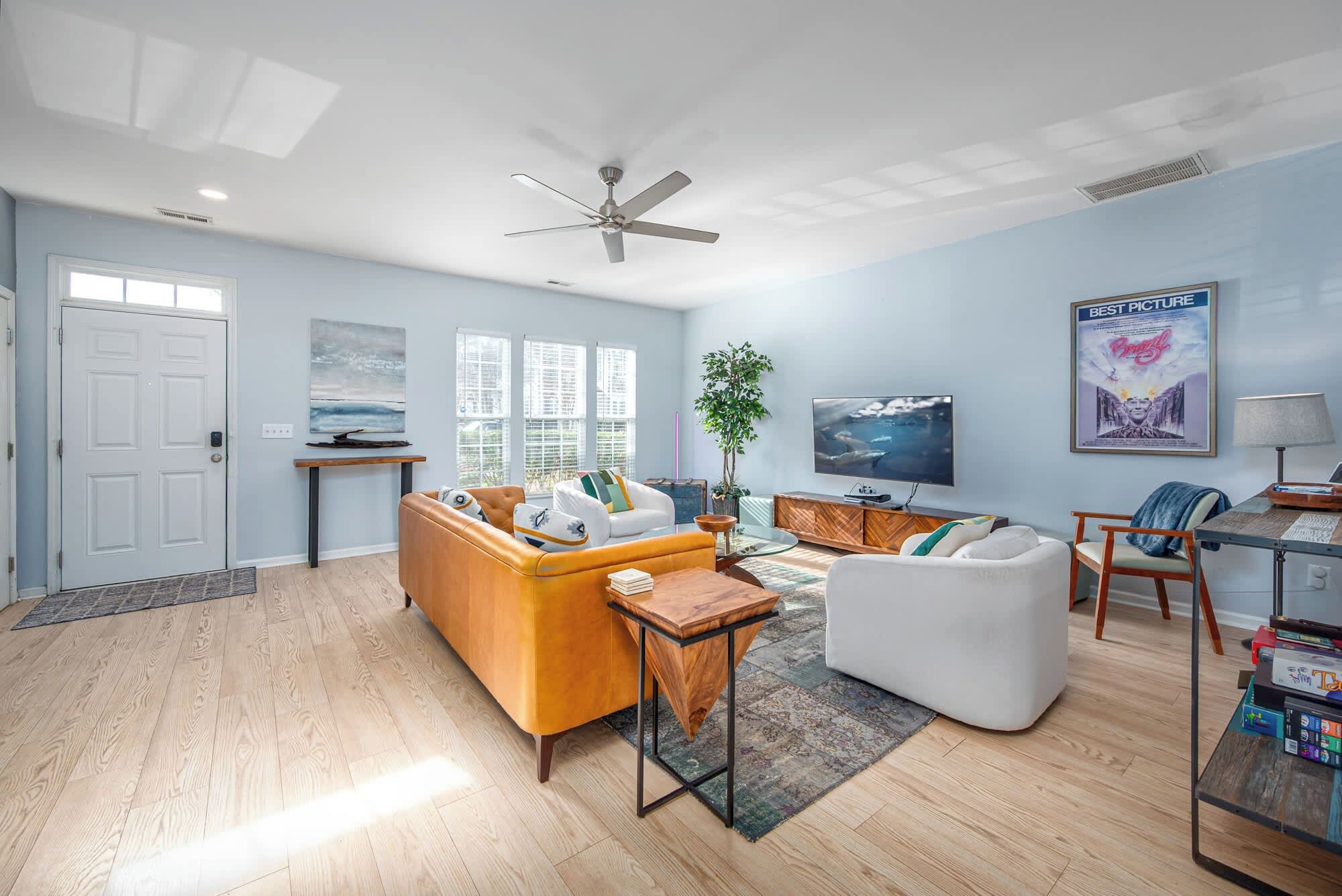 A modern living room with a sofa, armchair, and coffee table. There is a television, a ceiling fan, and a poster on the wall.