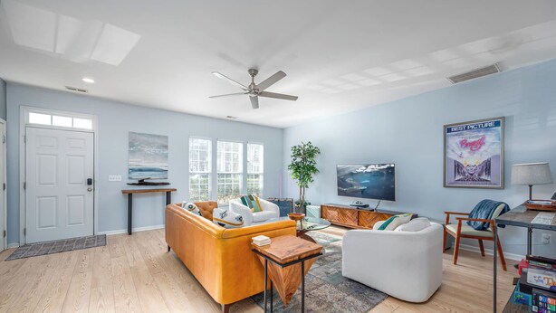 A modern living room with a sofa, armchair, and coffee table. There is a television, a ceiling fan, and a poster on the wall. - Modern 3-br Townhome w Private Backyard Near RTP (Morrisville)