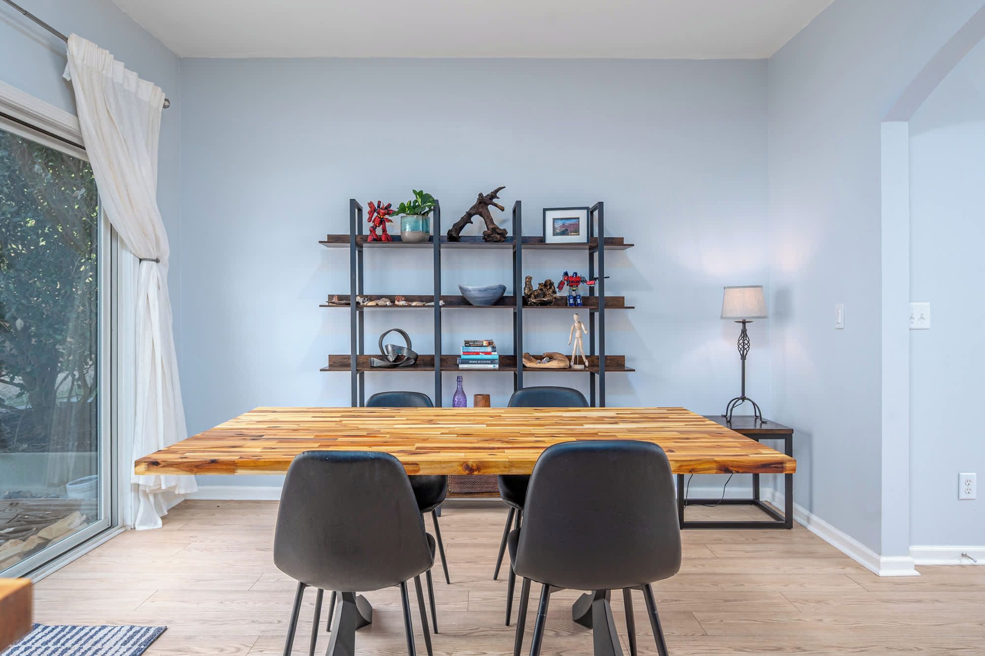 Ein Essbereich mit einem Holztisch, vier schwarzen Stühlen, einem Metallregal mit verschiedenen Gegenständen und einer Stehlampe.