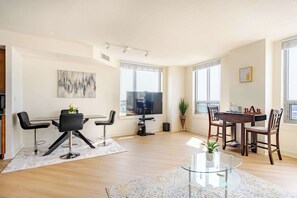 A modern living room with a dining area, large windows, a flat-screen TV, and a bar area.