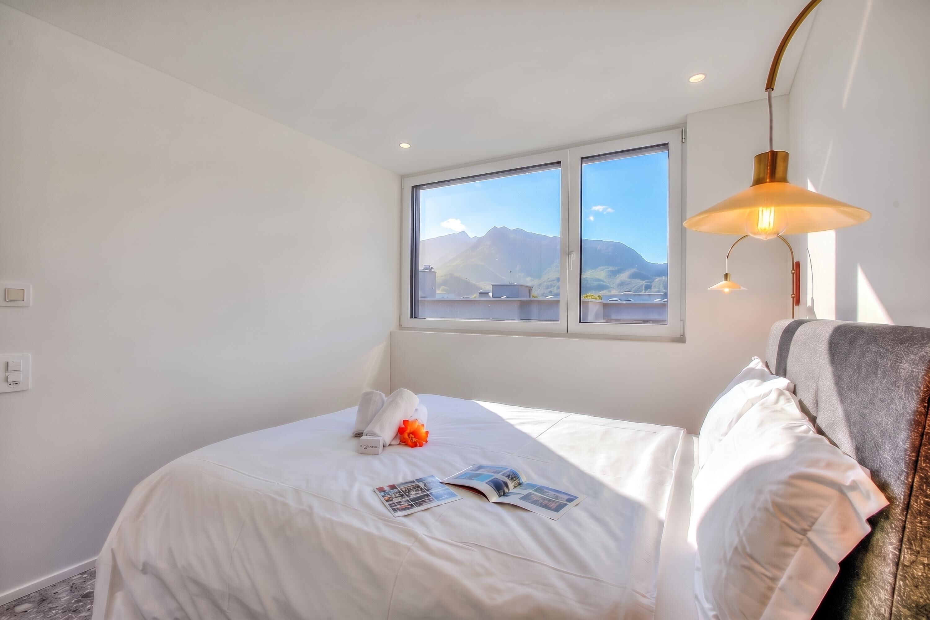A hotel room with a bed, a window with a mountain view, a lamp, and a power outlet.