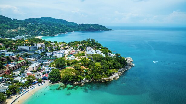 Vue aérienne d’une ville côtière avec une plage, des bâtiments et une chaîne de montagnes en arrière-plan.