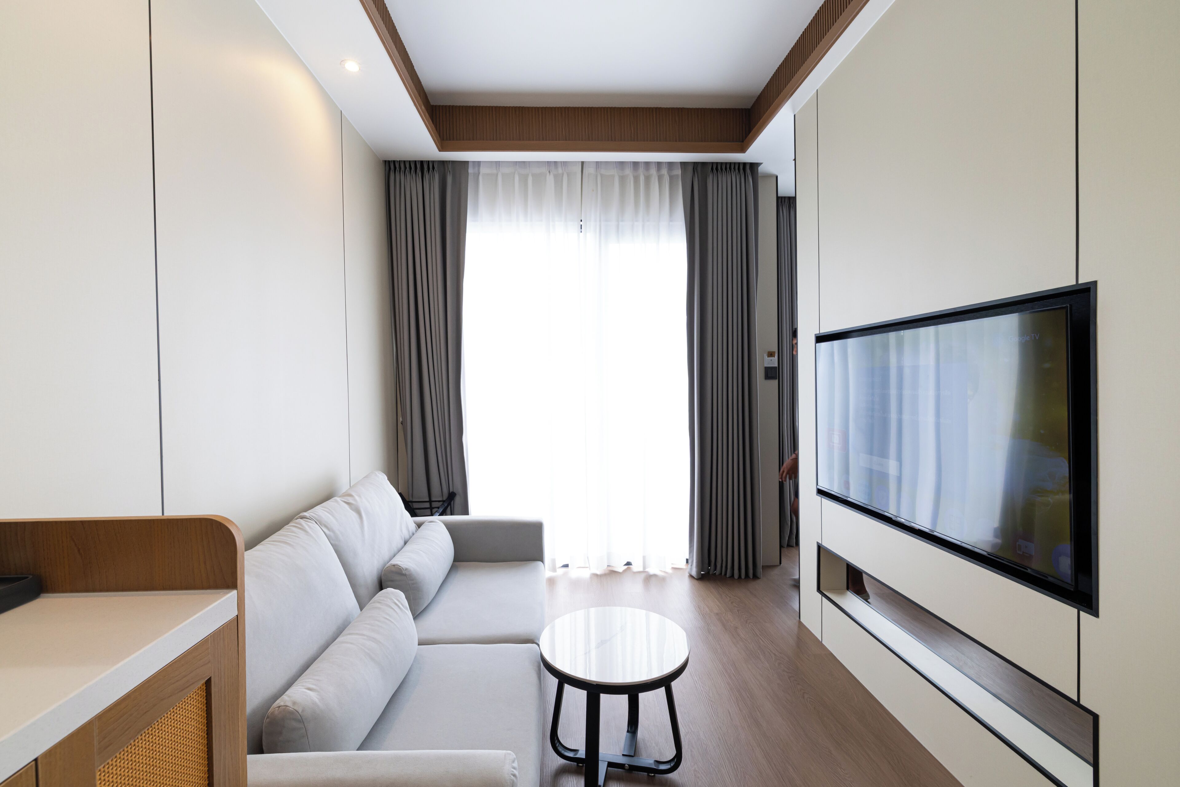 A modern hotel room with a sofa, a round table, a TV, and a large window with curtains.