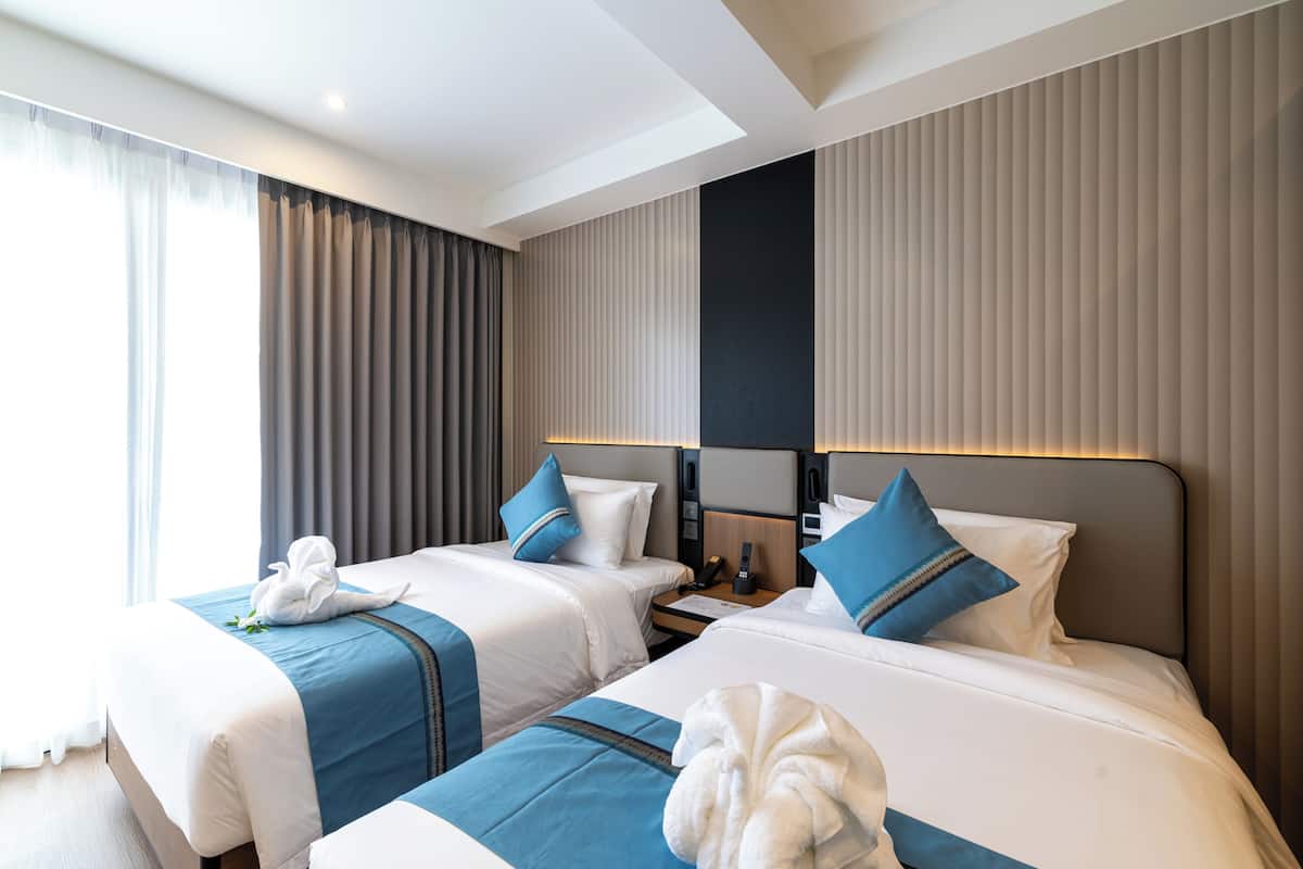 Habitación de hotel con dos camas, una mesita de noche, una lámpara y una ventana con cortinas.