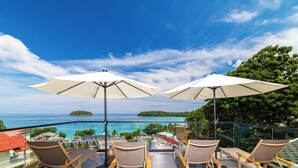Un balcon doté de quatre chaises longues et de deux parasols, offrant une vue sur une mer d’un bleu limpide et une petite île.