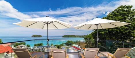 A balcony with four lounge chairs and two umbrellas, overlooking a clear blue sea and a small island.