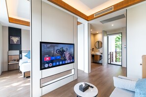 Moderni hotellihuone, jossa on taulutelevisio, sänky, sohva, pieni pöytä ja kylpyhuone, joka näkyy avoimesta ovesta.