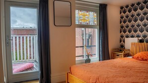Un dormitorio con cama, ventana con cortinas, una silla de mimbre y un radiador.