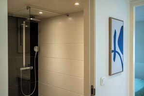 Un baño moderno con ducha envuelta en vidrio, una obra de arte abstracta enmarcada en la pared, y una puerta visible que da a otra habitación.