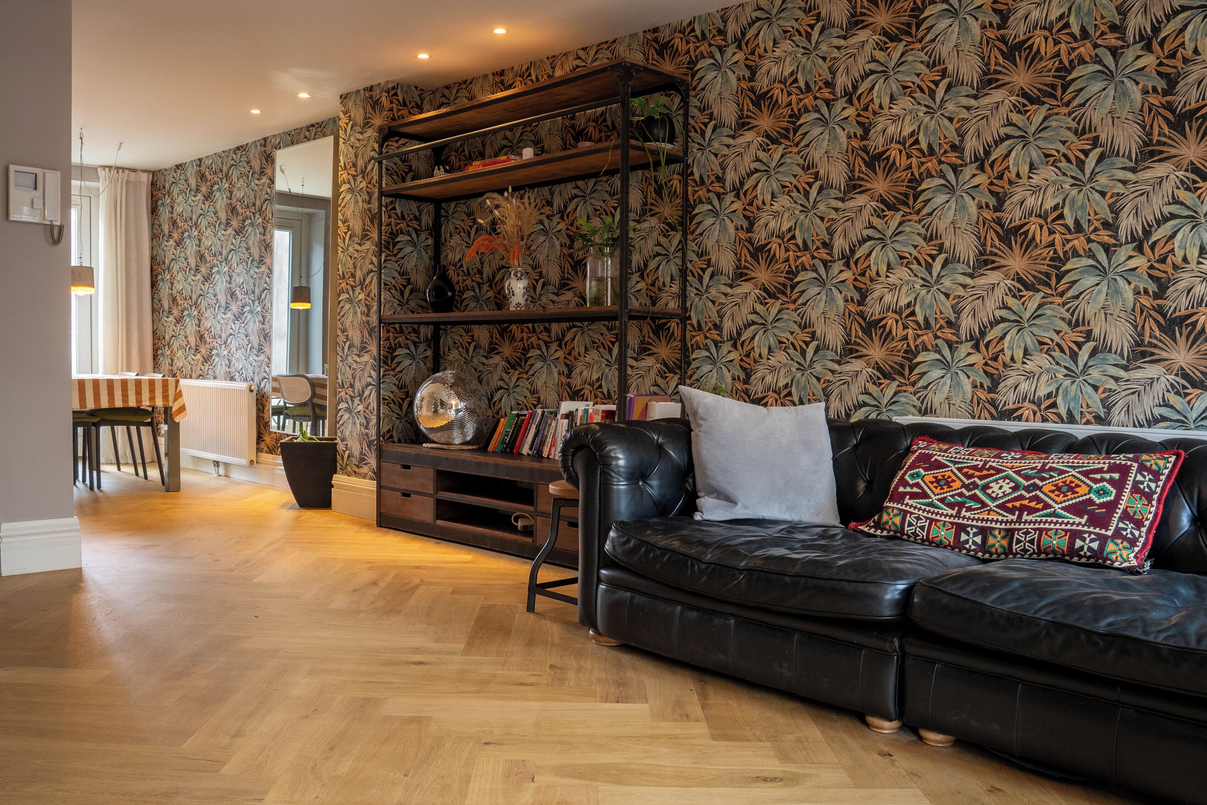 Een woonkamer met een zwarte leren bank, houten vloeren, een boekenkast en behang met een patroon.