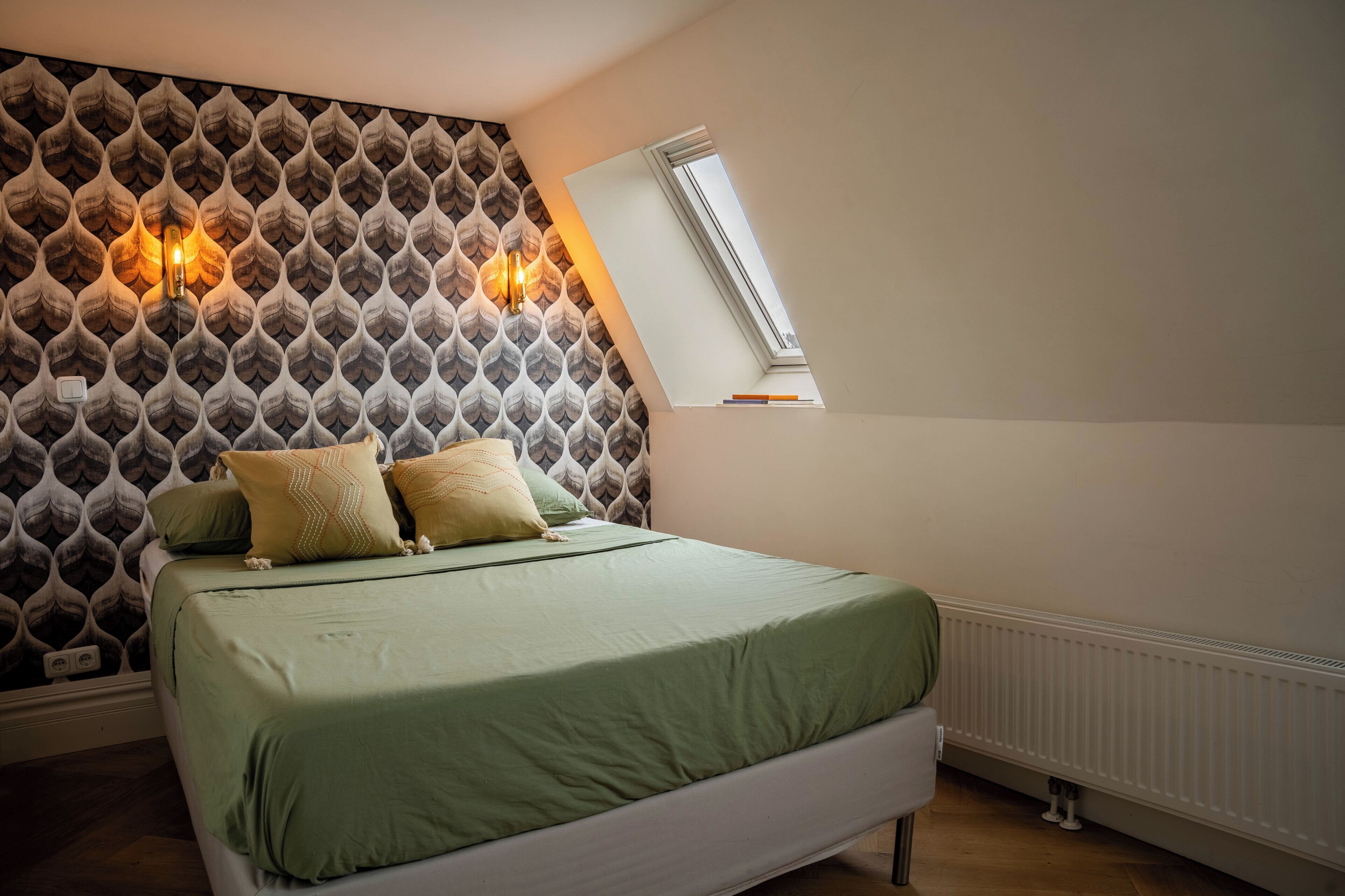 Een slaapkamer met behang met een patroon, een bed met groene beddengoed, twee wandlampen en een dakraam.