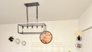 Ruokailutila, jossa on neljälle katettu pöytä, riippuvalaisin, kello ja ruukkukasvi.