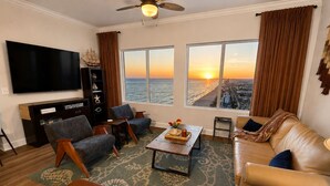 A living room with a large TV, two sofas, a coffee table, and a view of the sunset over the ocean. - Golden Hour Penthouse  -  Beach Front,  2 BR/2 Bath Penthouse (Panama City Beach)
