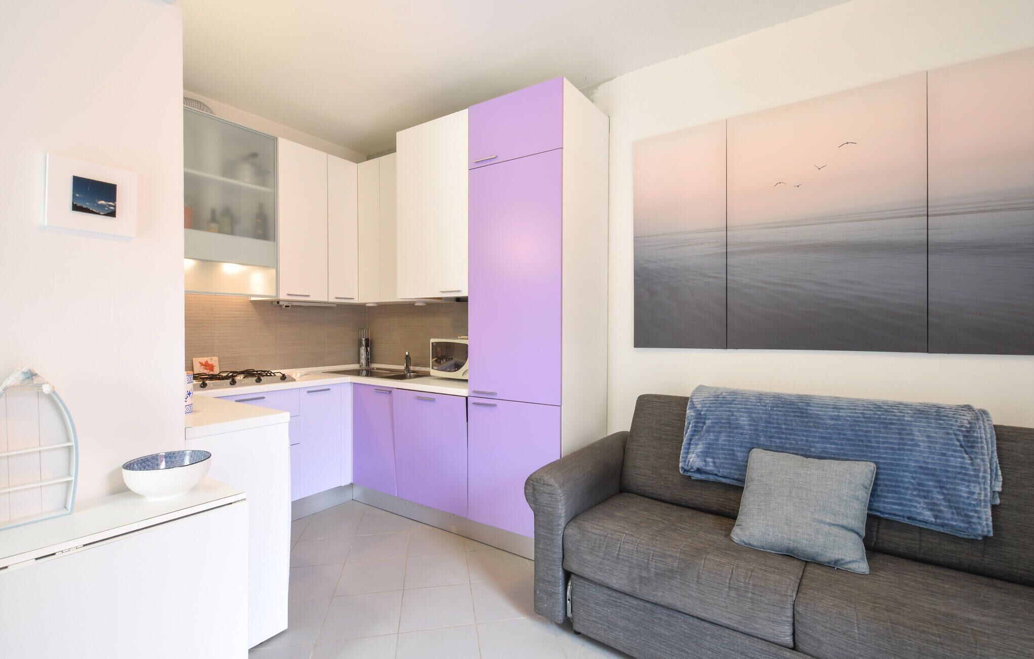 A compact kitchen with white and purple cabinets, a grey sofa with a blue blanket, and a large abstract painting on the wall.