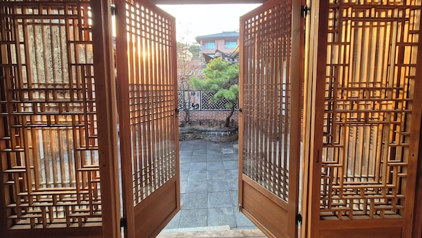Una puerta de madera con un diseño de celosía intrincado que da a un patio con un muro de ladrillo y un árbol.