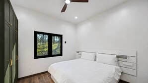Una camera da letto con un letto, una finestra vista foresta, una ventola a soffitto e pavimento in legno.