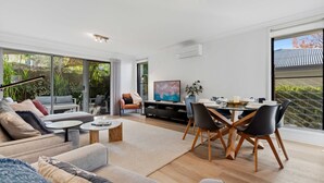 A modern living room with a sofa, coffee table, TV, and dining area with chairs and a table. - Coastal Cove by Experience Jervis Bay (Huskisson)