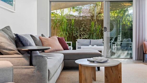 A modern living room with a grey sofa, a round wooden coffee table, and a glass door leading to an outdoor area with plants. - Coastal Cove by Experience Jervis Bay (Huskisson)