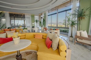 Ein großzügiger Wohnbereich mit einem gelben Ecksofa, einem runden Couchtisch und verschiedenen Sitzgelegenheiten, darunter eine Bank und Sessel. Große Fenster bieten einen Ausblick ins Freie.