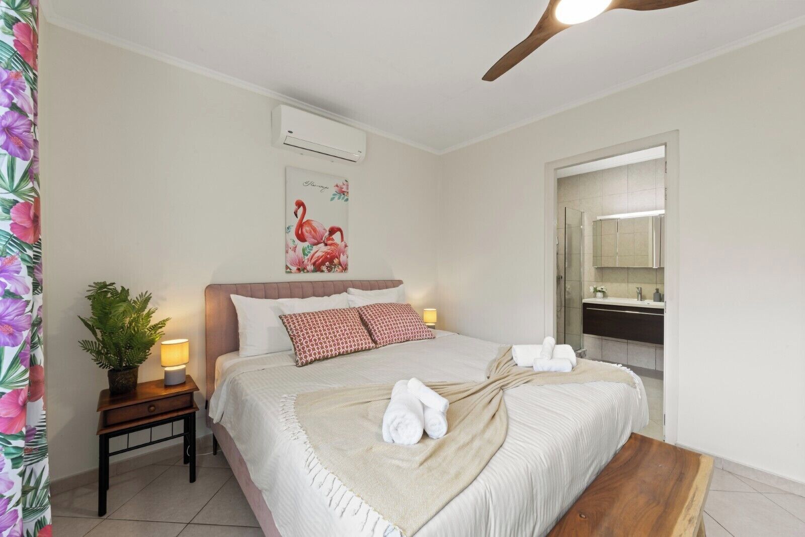 Un dormitorio con cama, mesita de noche, planta en maceta, decoración en la pared y un baño visible a través de una puerta abierta.