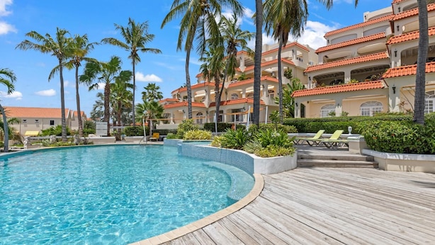 A swimming pool with a wooden deck, surrounded by palm trees and a multi-story building with red-tiled roofs. - Casa Piscadera · Ocean View · Pool · Beaches (Piscadera)